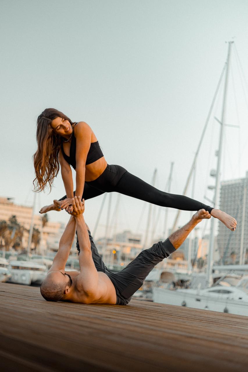 An acroyoga duo in a front-plank pose on a marina dock at sunset, sailboats behind them.